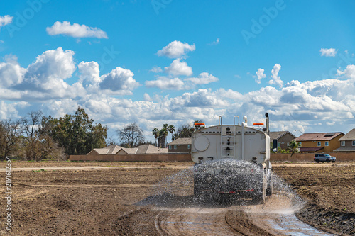 Water truck sprays dirt road at construction site, preparing ground for construction near residential area.
