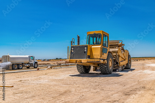 Heavy earthmoving scraper parked next to water tanker that spraying road surface at construction
