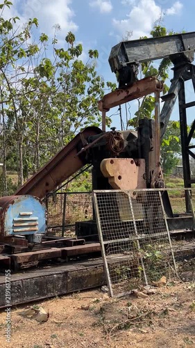 Rusty oil pump jack in countryside with green trees, suitable for industrial, machinery, energyrelated, and ruralthemed projects.
