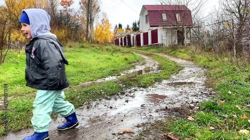 Cute little toddler child kid boy playing outside in boots in muddy puddle after rain in garden,home backyard at autumn. Carefree childhood. Moody fall weather forecast