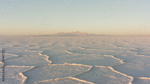 A vast salt flat landscape with hexagonal cracked patterns on the ground stretching towards distant mountains under a soft, clear sky during the golden hour, creating a serene and desolate atmosphere