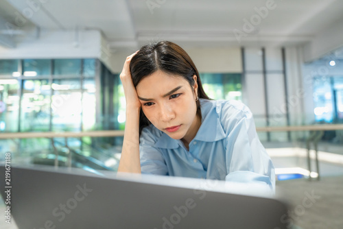Young Asian businesswoman frowning with hand on her head while using AI software on a laptop, feeling overwhelmed by new technology at a modern office desk.
