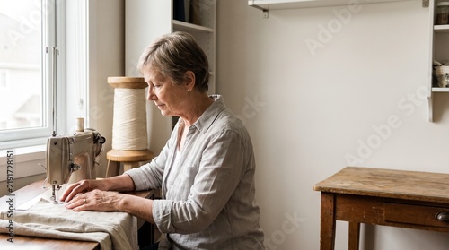 Senior woman focused on sewing fabric with a vintage machine in a bright sunlit workshop with wooden furniture and neutral decor