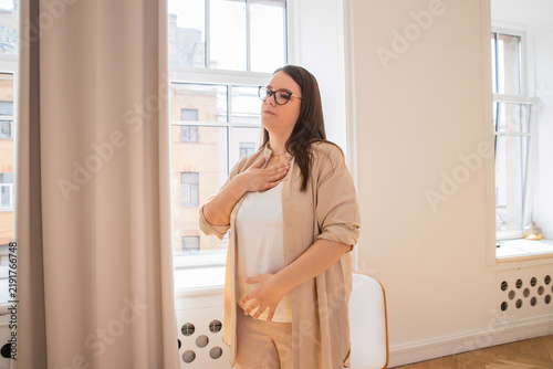 Woman psychologist practicing diaphragmatic breathing exercise with hand on chest and belly. Calm therapy session in bright room near window.