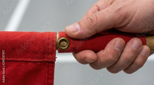 Close up of a hand holding a wooden pole with a red fabric flag attached via a metal grommet