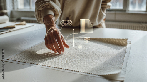 Hand reaches toward beige textile fabric laid flat on a white table within a bright studio. Glowing line art icons of yarn and needle float above the woven material in soft sunlight.