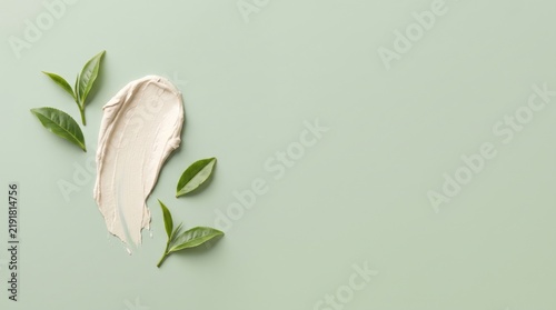 Minimalist background with a smear of light beige cosmetic cream and fresh green tea leaves arranged on a soft pastel sage green surface