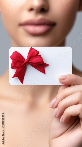 White gift card with a voluminous red satin bow held in a person's hand against a light background. Gift, shopping, holiday, and thoughtful present concept.