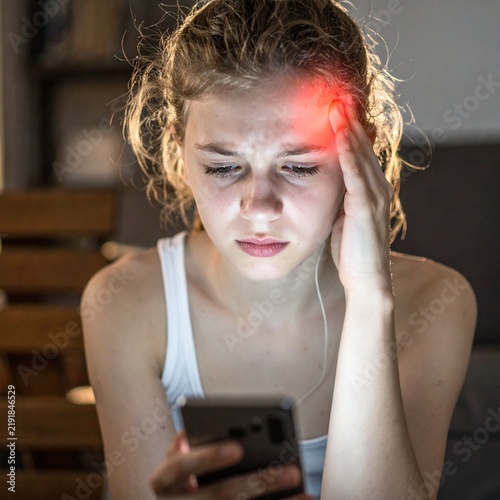 Stressed young woman experiencing a severe headache while looking at her glowing mobile phone at night.
