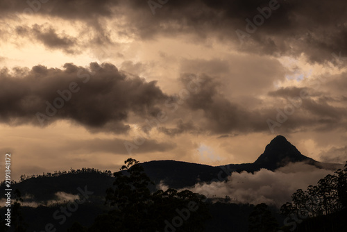 A distinctive mountain peak rises above misty hills beneath dramatic clouds in the central highlands of Sri Lanka near Maskeliya.