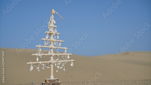 Driftwood and shell Christmas tree standing on sandy ground with desert dunes and clear blue sky in the background, representing sustainable holiday travel