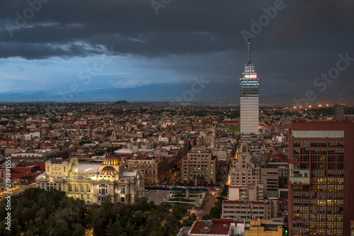 Panoramic view of the historic center of Mexico City during twilight. The illuminated Palacio de Bellas Artes and Torre Latinoamericana stand out against a dramatic cloudy evening sky and distant moun