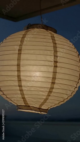 Vertical video: Swaying paper lantern on porch at dusk, breeze causing spin, internal LED glowing