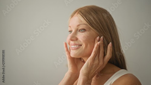 Young blonde woman massaging cheeks and jawline with hands, smiling while applying skincare in bright studio; self care calm.