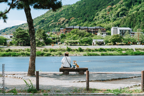 Peaceful riverside scene in Arashiyama, Kyoto, Japan, showing a person sitting on a bench with a Shiba Inu dog overlooking the Katsura River. Calm spring landscape combining nature, companionship.