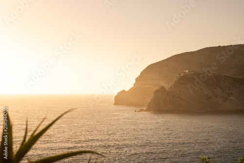 View of the rocky coastline of Ischia island at sunset with calm sea and golden light in Ischia, Campania, Italy.