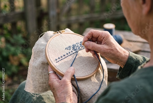 Senior Woman Hands Embroidering Blue Pattern on Linen Fabric with Wooden Hoop Outdoors