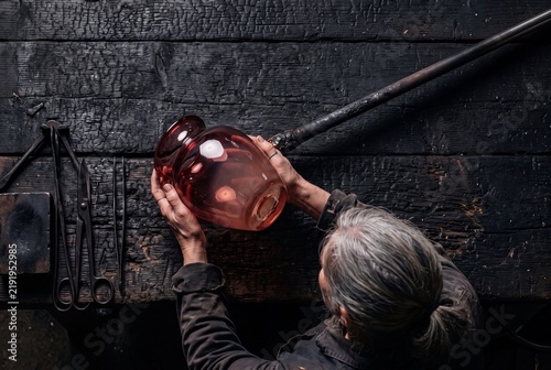 Glassblower Shaping and Inspecting Handcrafted Glass Vase at Charred Wooden Workshop Table