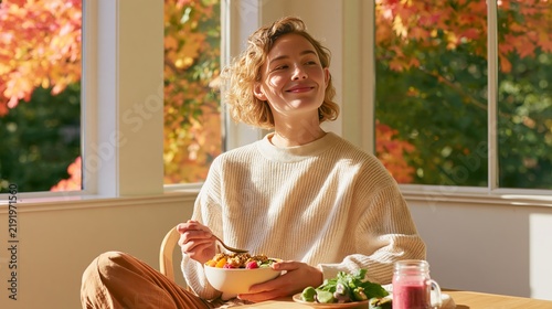 Young woman enjoying a smoothie bowl while sitting at a wooden table in a cozy room with large windows and autumn foliage visible outside