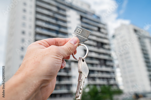 Hand Holding House Keys with Metal House Keychain in Front of High Rise Apartment Building Exterior