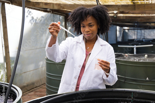 Woman scientist wearing white lab coat holding pipette and beaker sampling water at greenhouse tank