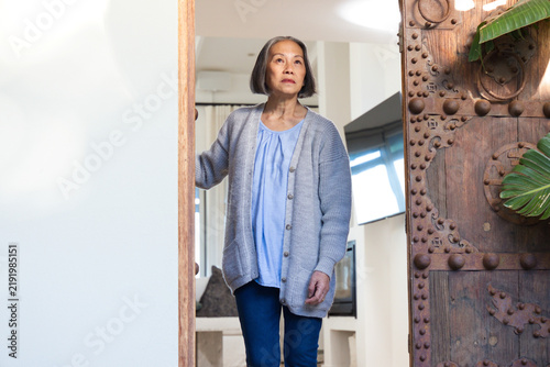 Senior Asian woman standing in doorway at home wearing gray cardigan and jeans, holding carved door
