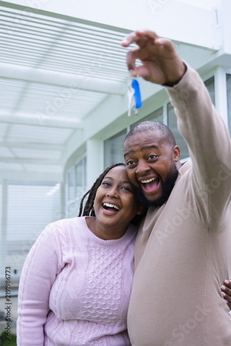 African American couple hugging, holding keys with blue fob under pergola on porch wearing sweaters