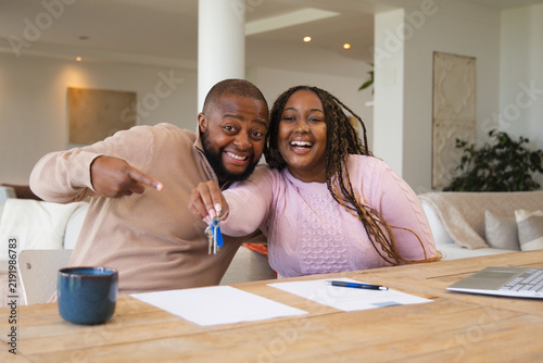 African American couple smiling, holding keys and pointing at signed papers at home on wooden table