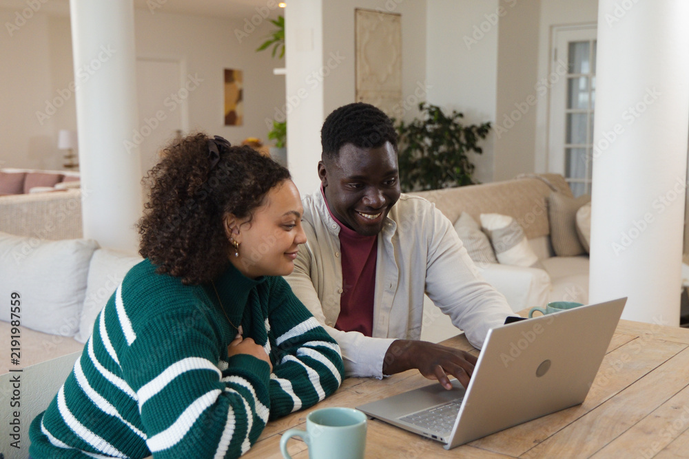 Obraz premium Couple sitting at wooden table at home, leaning and pointing at silver laptop with mugs