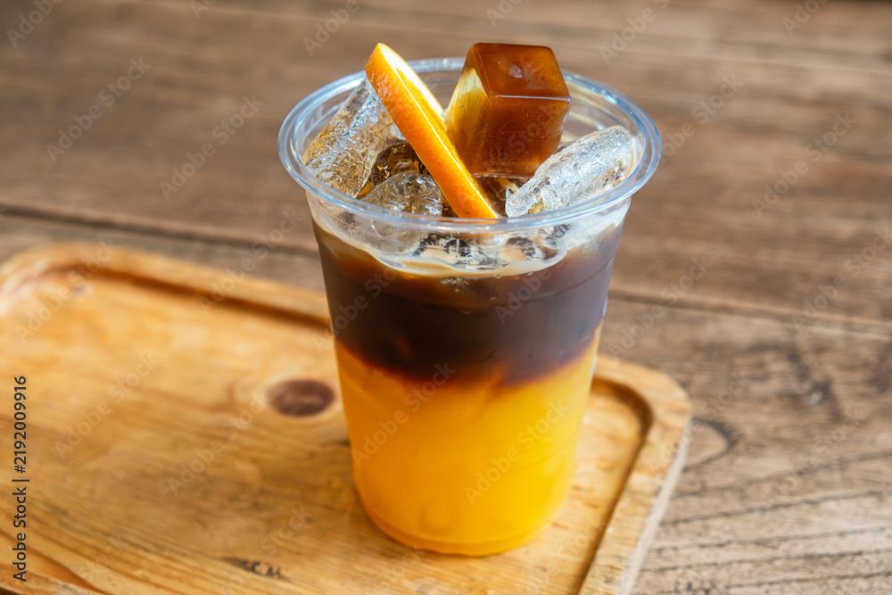 Obraz premium Cold Bumble coffee combining fresh orange juice and espresso with ice, orange slice and coffee ice cube, served on wooden tray in Thailand. Coffee recipe, specialty cafe menu, tropical refreshment