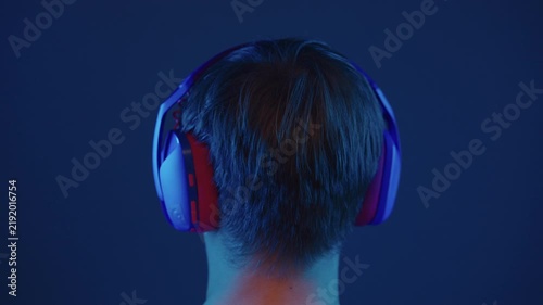 Back view of young person wearing colorful headphones in dark room with blue red neon light, ad moves slightly, listening to music playing video game. Immersive digital entertainment concept.
