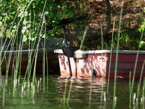 Fotografie Small Red Fishing Boat with Electric Trolling Motor Moored Behind Reeds