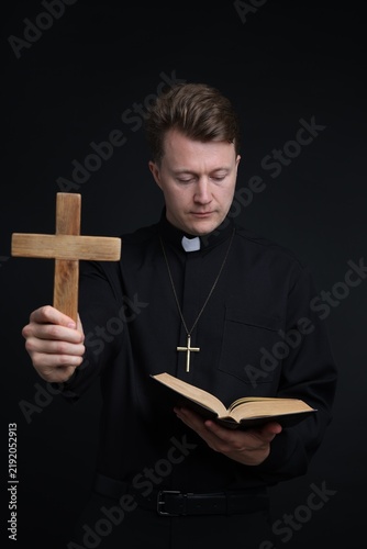 Foto Priest in cassock with wooden cross reading Holy Bible on black background