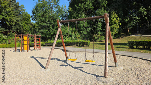 Wooden swing set with yellow seats standing on a gravel outdoor playground surrounded by green trees
