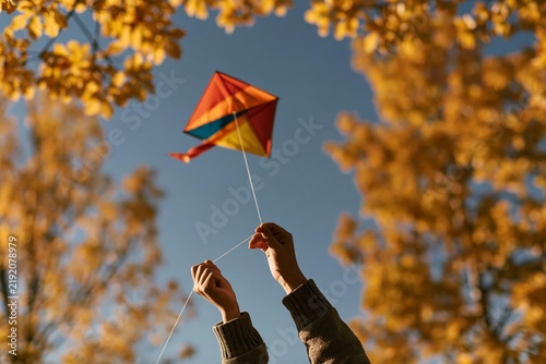 Child holding string of colorful diamond kite against blue sky and autumn leaves