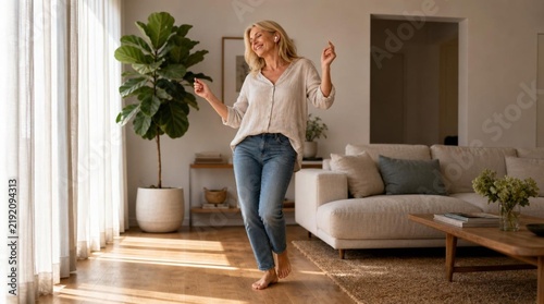 Carefree mature blonde woman in cream linen blouse and blue jeans dances barefoot on wooden floor of bright modern living room with large fiddle leaf fig plant and neutral sofa, joyful home lifestyle