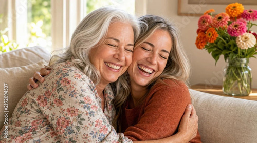 Close-up of joyful silver-haired mother and adult daughter embracing and laughing together on a beige linen couch in sunlit living room with fresh zinnia bouquet