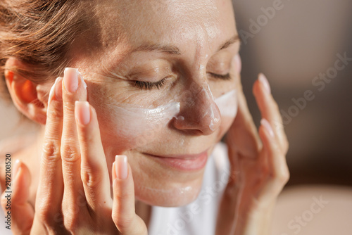 Foto Mature Woman Applying Face Moisturizer in Warm Morning Sunlight
