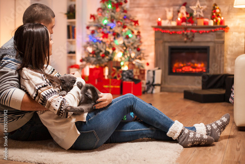 Foto Relaxed couple cuddles on the floor while the woman holds a gray cat, surrounded by a Christmas tree, gifts and a glowing fireplace in a cozy holiday home