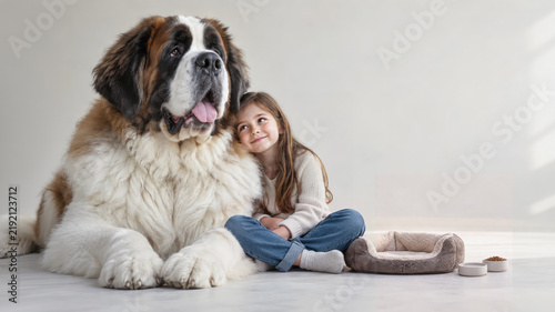 A smiling girl sits leaning against an oversized Saint Bernard beside a tiny dog bed and bowls, creating a whimsical contrast in size and a warm portrait of companionship
