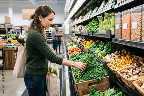 Woman choosing fresh organic kale from produce shelf with reusable tote bag in supermarket aisle