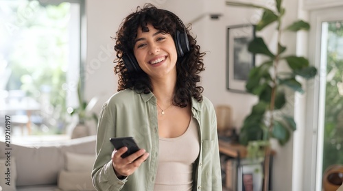 Cheerful young woman with curly dark hair smiling brightly while wearing over-ear black headphones and holding a smartphone in a cozy modern living room interior with plants