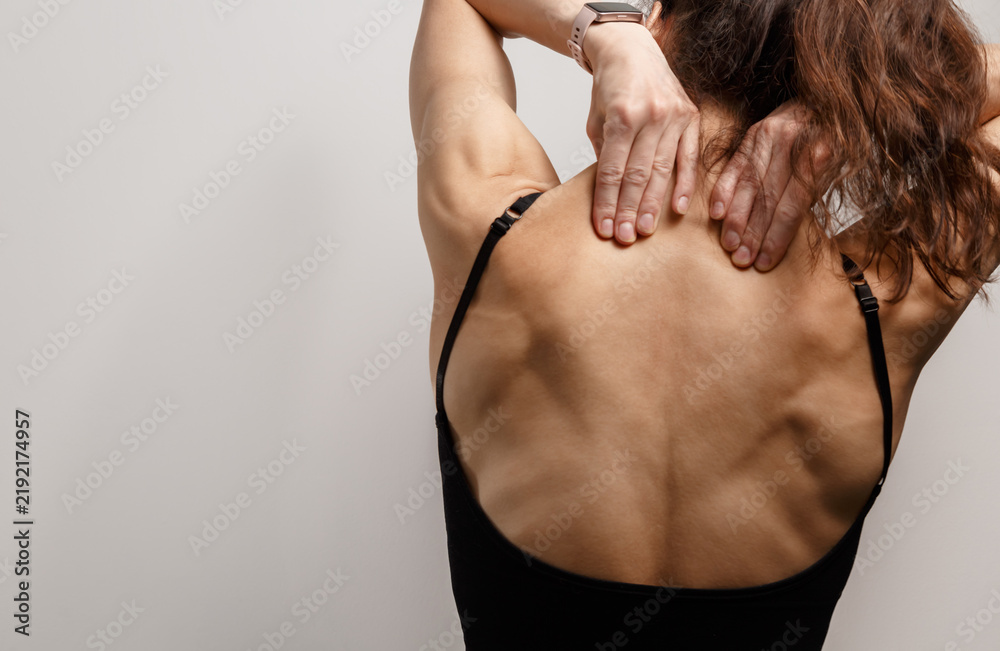 Obraz premium Athletic woman herself hands massaging her upper back, neck, trapezius muscles in pain, tension, soreness, recovery, posture, wellness. Selfcare after exercise with studio light copy space. Rear view