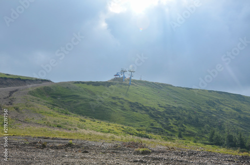Chairlift station sits atop a green mountain ridge under dramatic sunlight breaking through dark clouds. Dragobrat, Carpathian Mountains, Ukraine