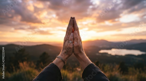 Hands held in prayer position at sunrise over a mountain landscape.