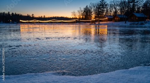 Winter evening scene with illuminated string lights over a frozen lake and cabins