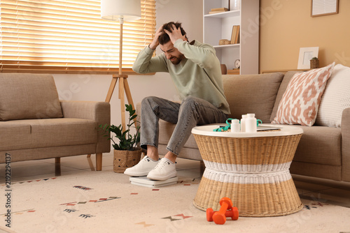 Stressed young man with weight scales sitting on sofa at home