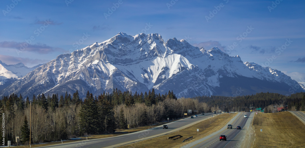 Obraz premium Highway traffic driving toward massive snow capped Mount Lougheed under clear blue sky in Canadian Rockies