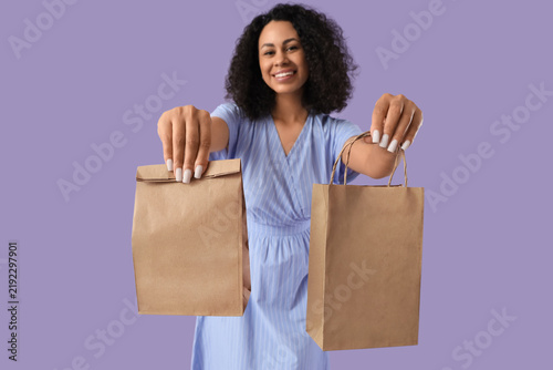 Young African-American woman with paper bags on lilac background. Food delive...