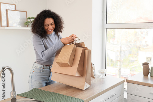 Young African-American woman putting paper bags on counter in kitchen. Food d...